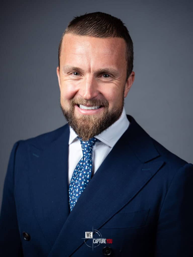 Professional corporate headshot of a smiling man in a navy suit and blue tie.