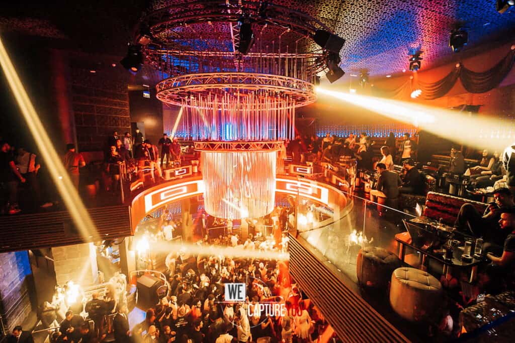 Crowded nightclub with colorful lights, DJ booth, and dancing crowd.