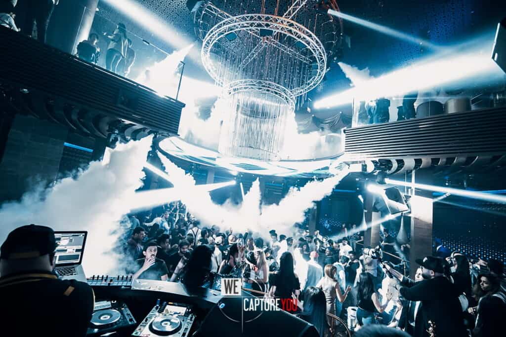 Crowd dancing in a vibrant nightclub with colorful lights and DJ booth.