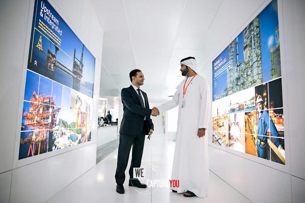 Two businessmen shaking hands in a contemporary office corridor with digital displays.