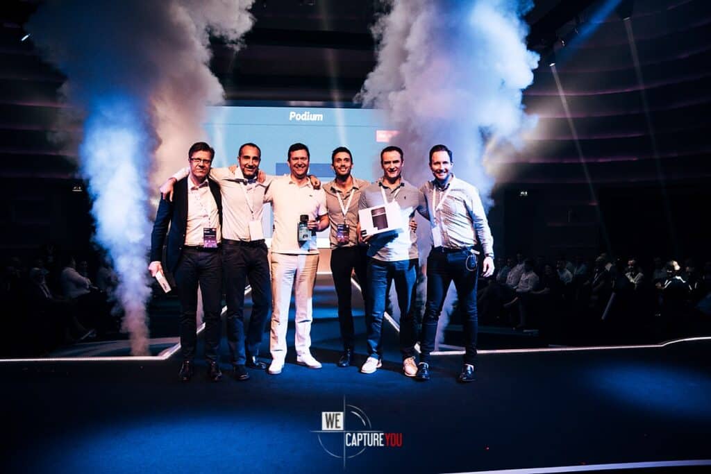 Group of six men on stage at a conference with smoke effects and a podium.