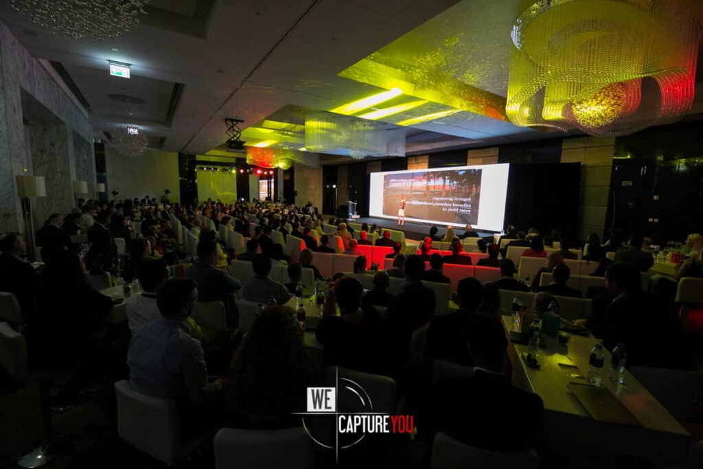 Large audience attending a conference with a speaker on stage at WeCaptureYou event.