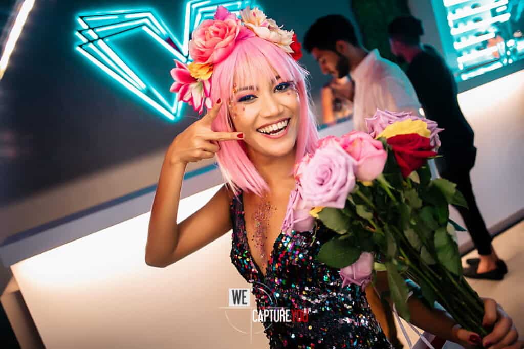 Smiling woman with pink hair and flower crown holding a bouquet of roses.