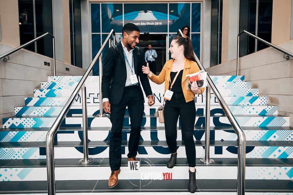 Two diverse professionals smiling and talking on conference stairs at WeCapture event.