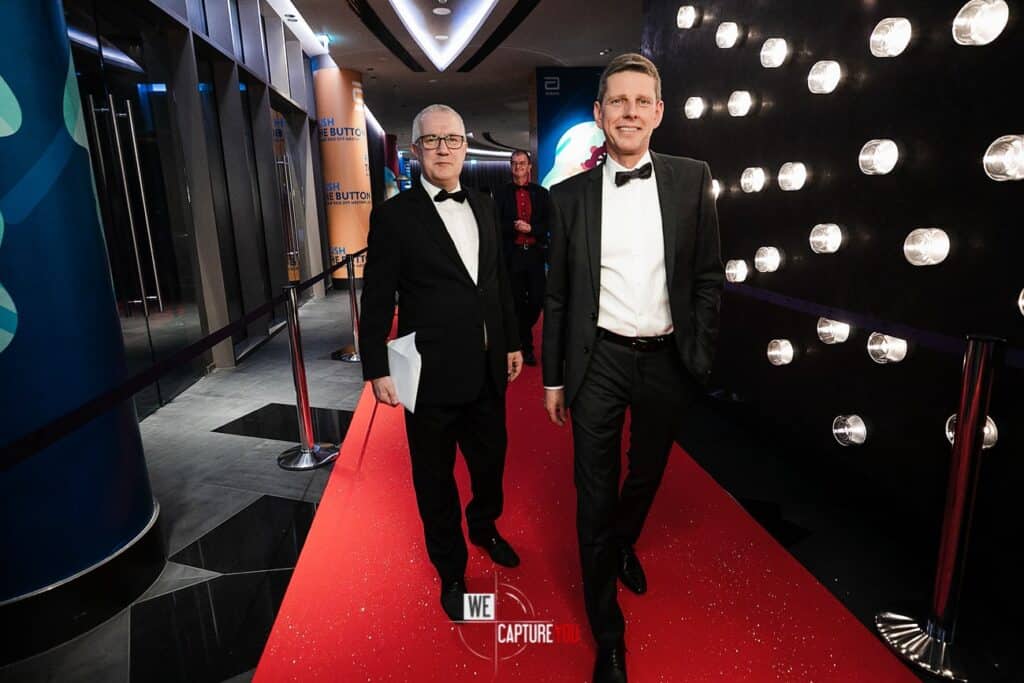 Two men in tuxedos walking on a red carpet at a formal event.