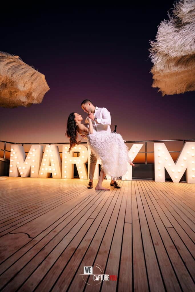 Couple dancing on a wooden deck with sunset sky and illuminated letters in background.