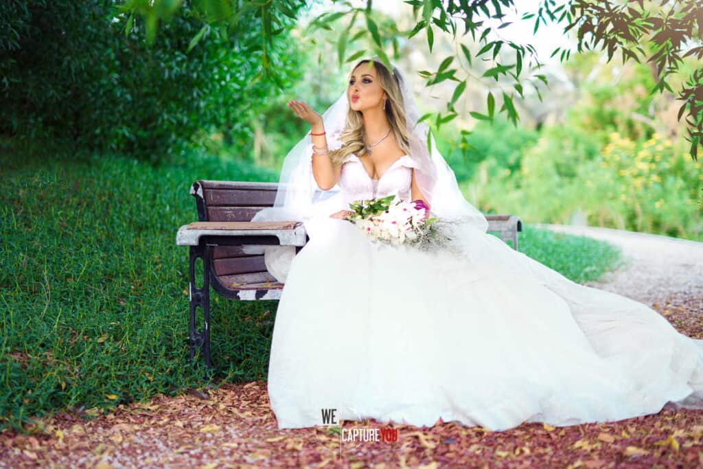 Beautiful bride in white wedding dress with bouquet on park bench.