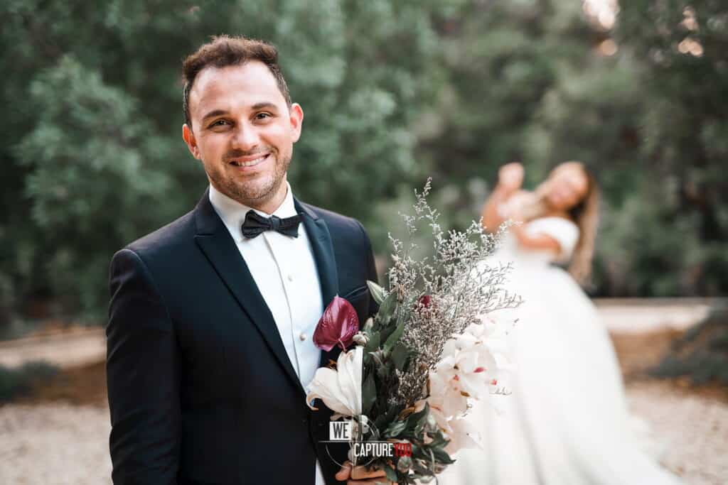 Groom holding a bouquet at wedding, smiling, outdoor setting.