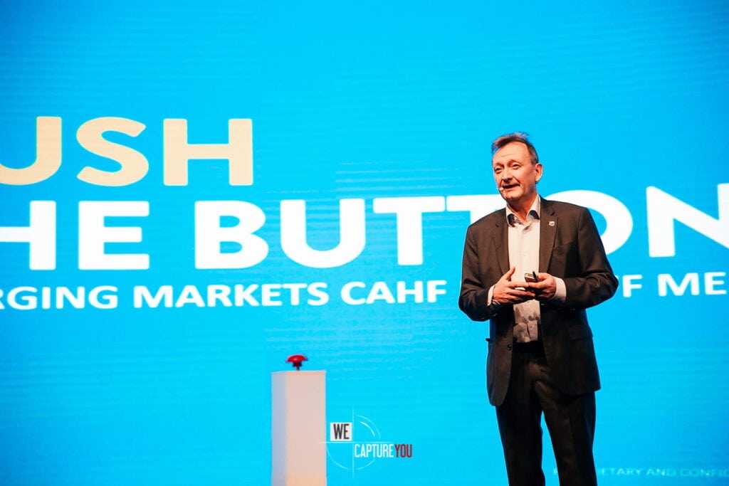 Businessman speaking at WeCapture conference with large screen background.