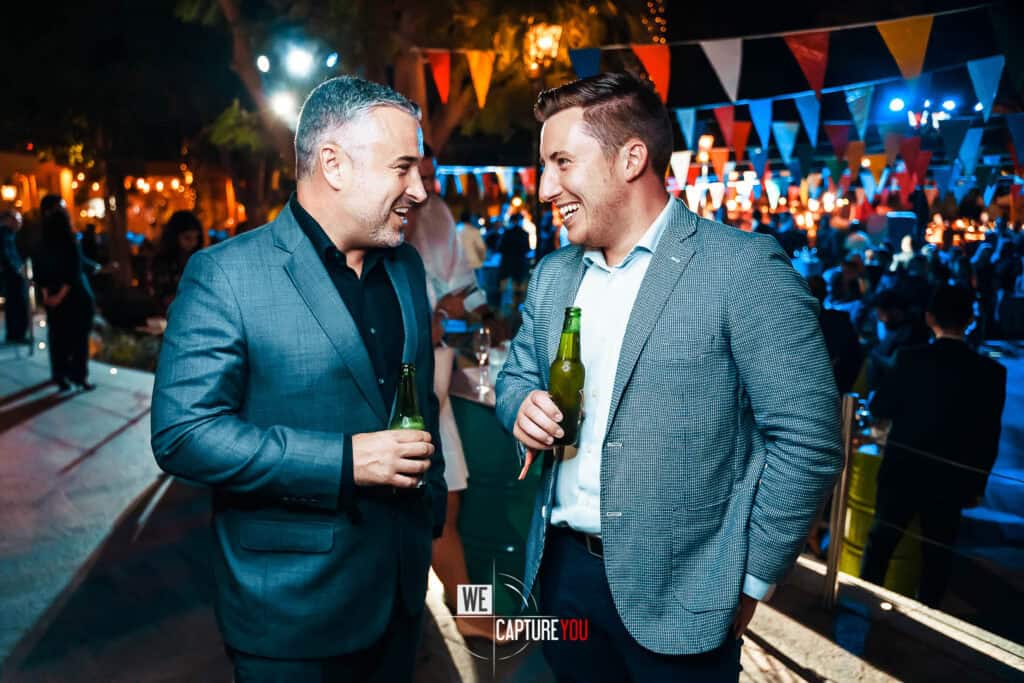 Two men in suits talking and drinking beer at outdoor party.