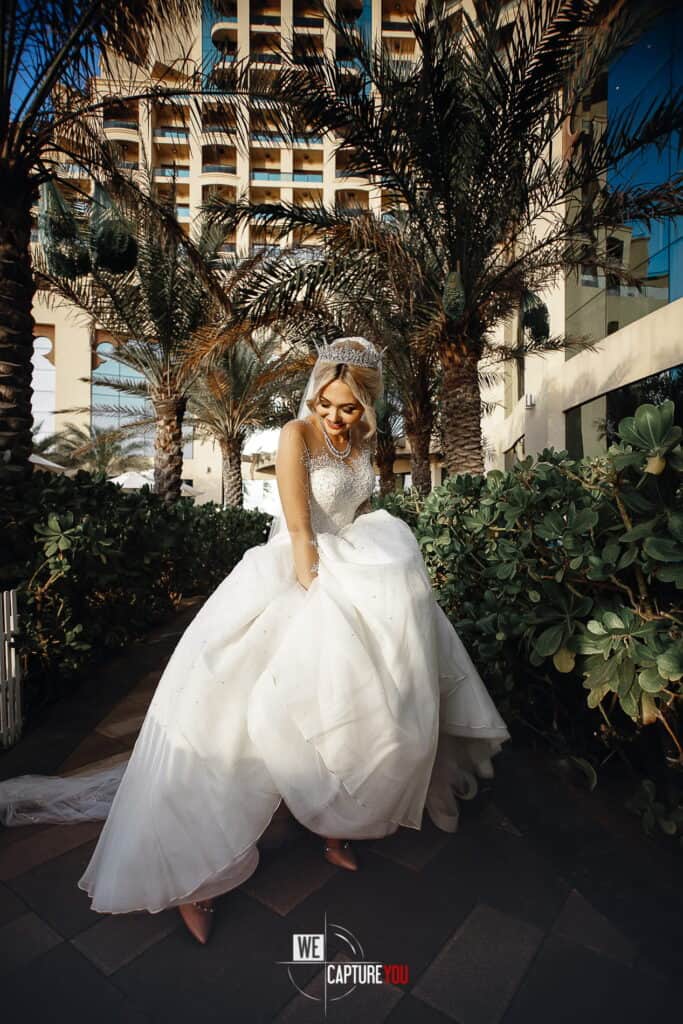 Beautiful bride in white wedding gown outdoors with palm trees and city buildings.