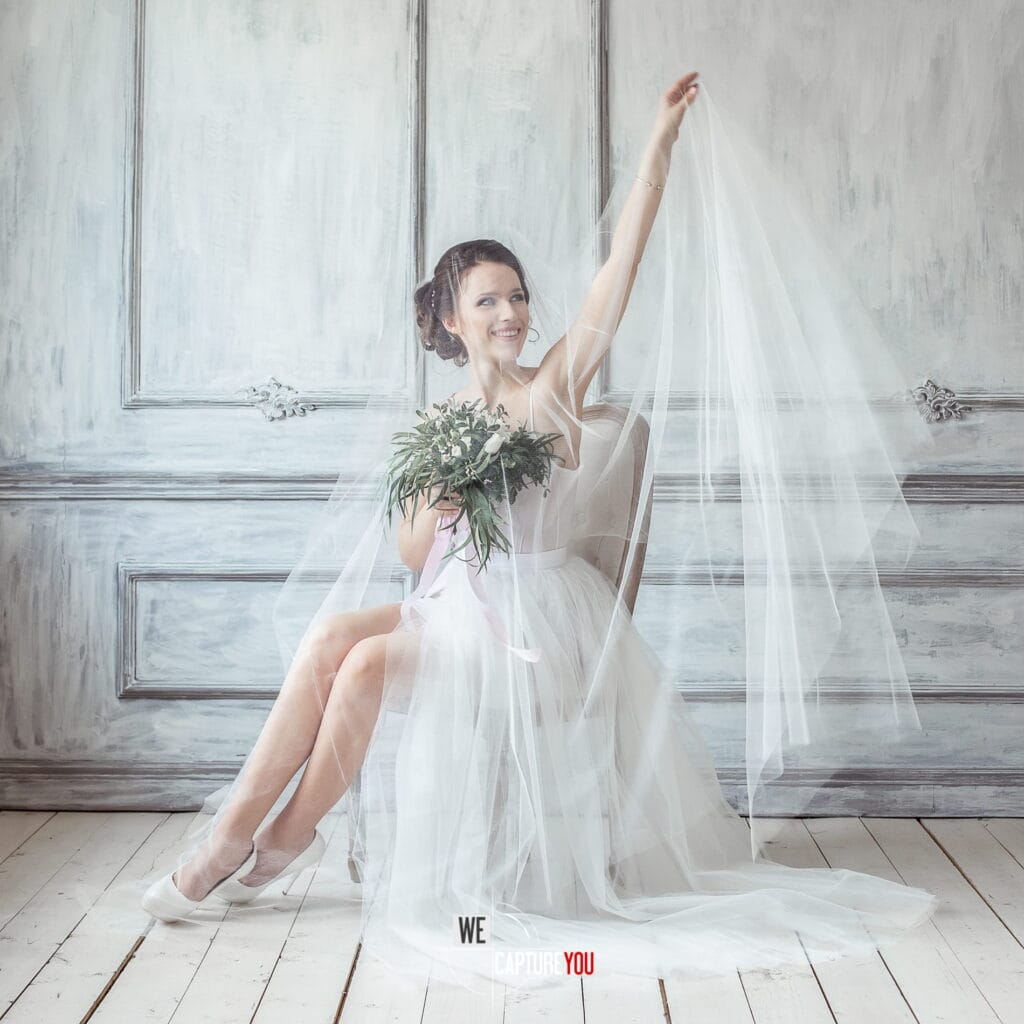 Beautiful bride holding bouquet and lifting veil in a stylish studio setting.