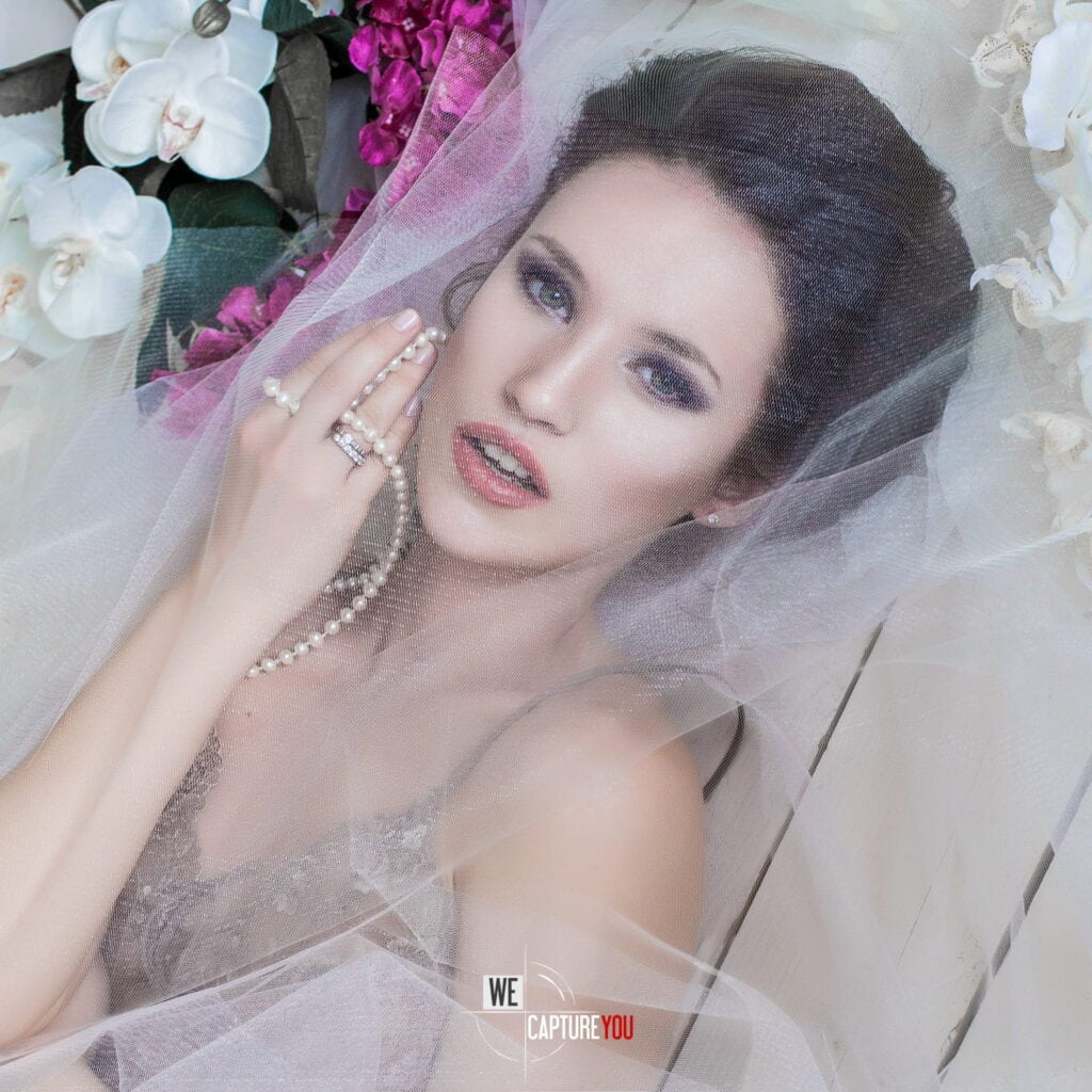 Beautiful bride with makeup, jewelry, and veil surrounded by flowers.