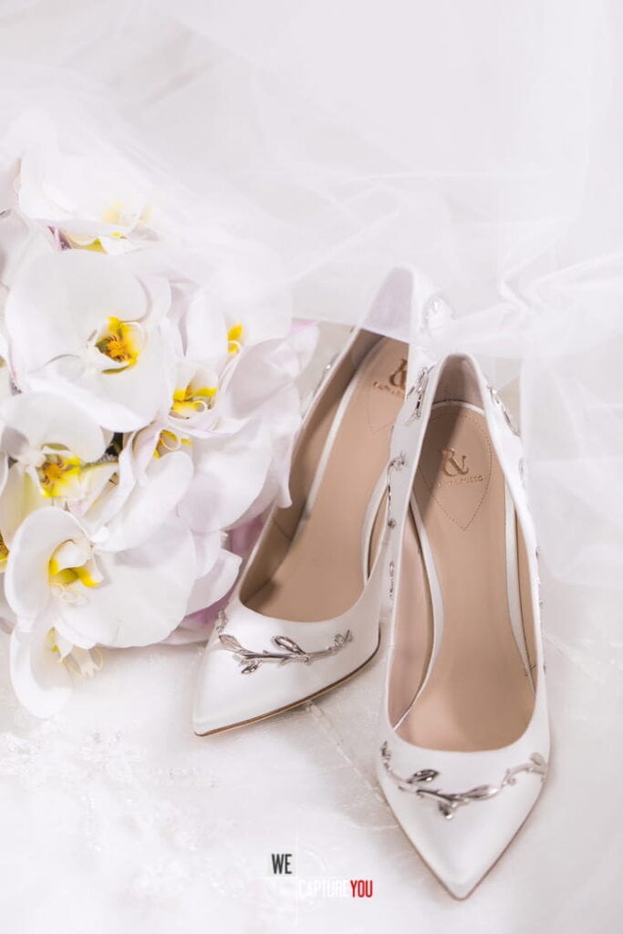 White high-heeled wedding shoes with floral embellishments on a soft white fabric background.