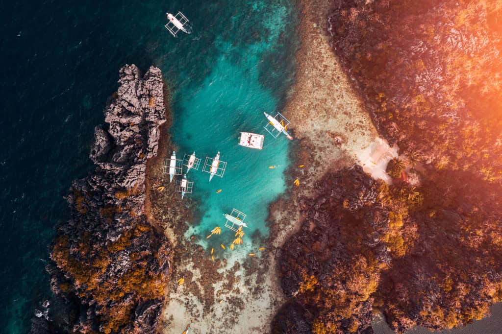 Drone aerial shot of boats near rocky coast with turquoise water and autumn foliage.
