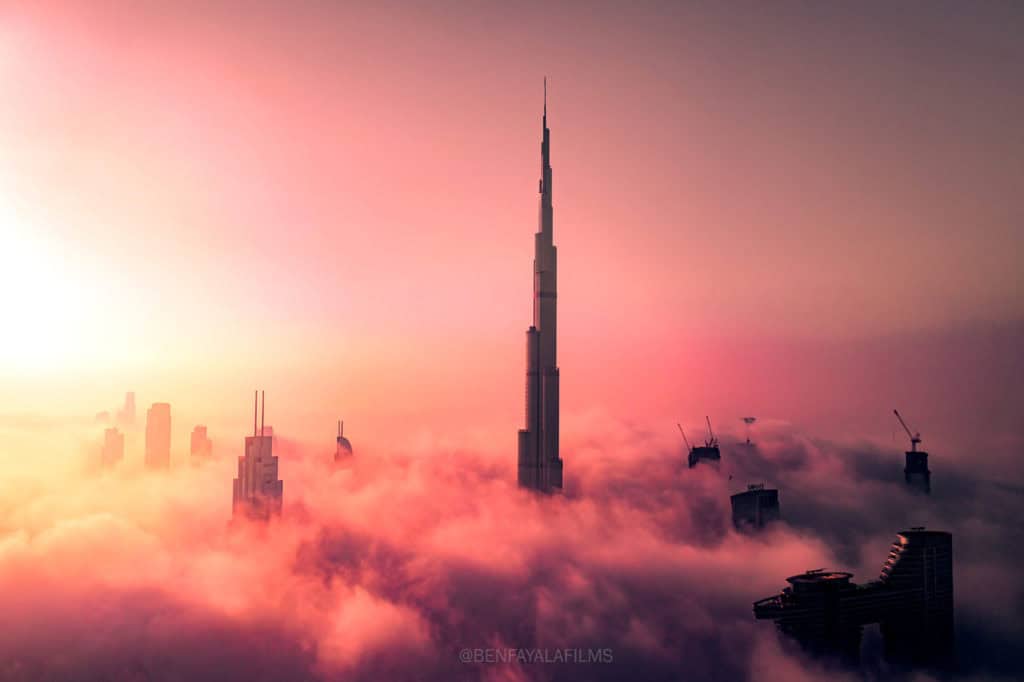 Aerial view of Dubai skyline with Burj Khalifa above clouds at sunrise.