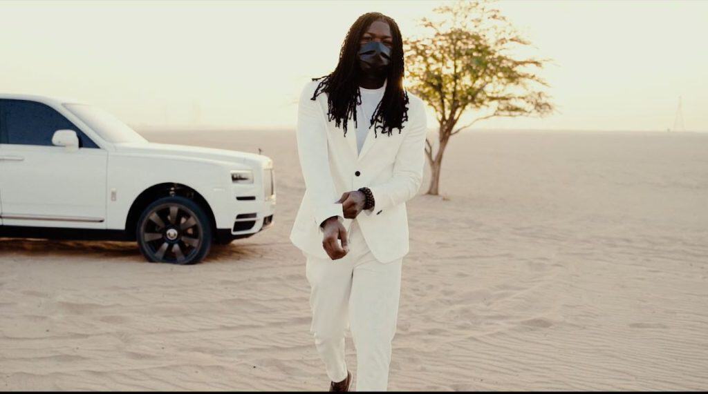 Stylish man in white suit walking on desert with luxury car in background.
