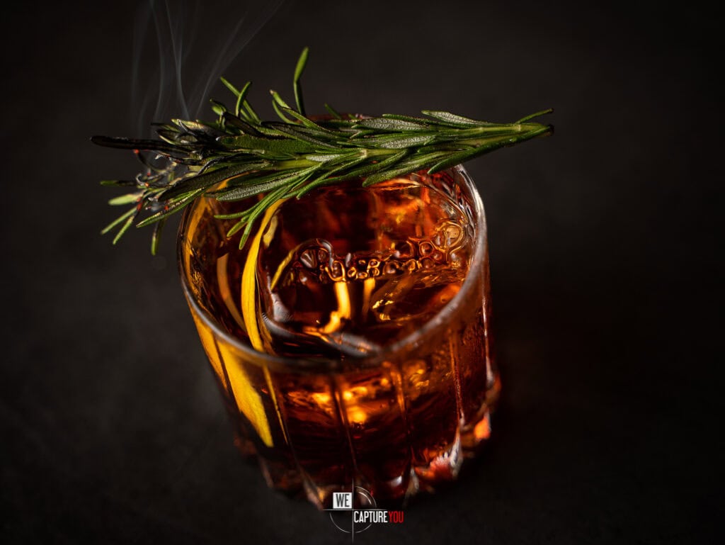 Glass of whiskey with ice, lemon slice, and rosemary sprig on dark background.
