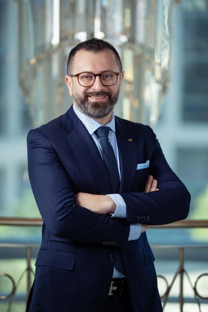 Professional corporate headshot of a smiling man in a navy suit and glasses.