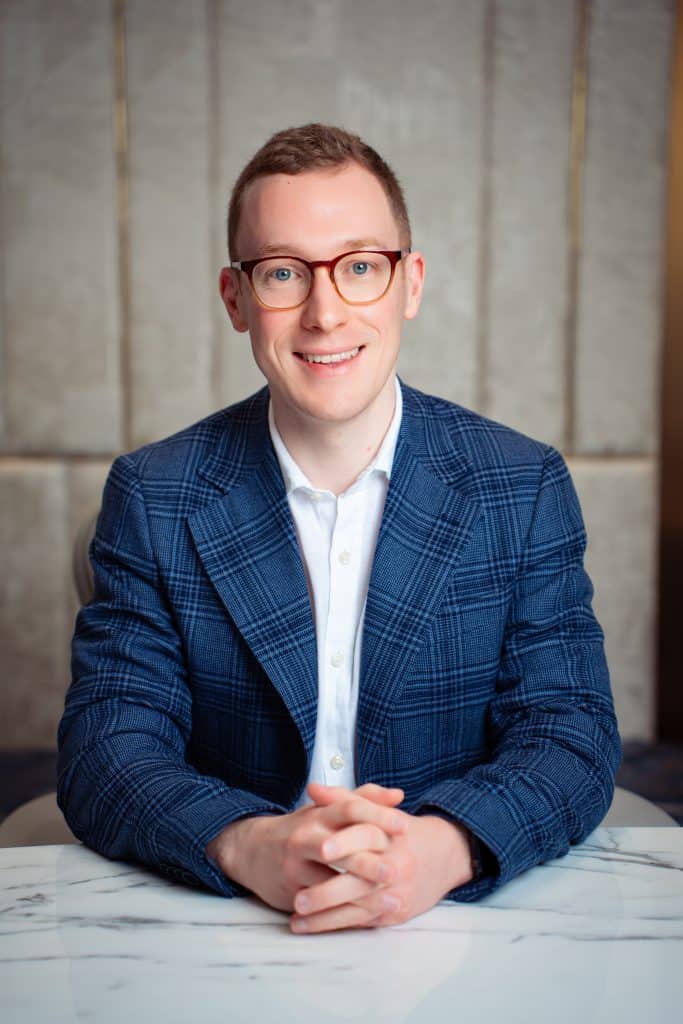 Professional corporate headshot of a smiling man in glasses and a blue blazer.
